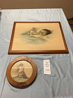 Photo showing both Bessie Pease Gutmann wall art prints on blue cloth. The larger rectangular piece 'Awakening' features a child holding a crown and has a simple wooden frame. The smaller round piece 'In Disgrace' shows a child with a dog standing in a corner inside a round wooden frame.