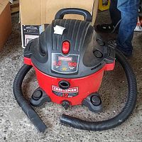 Photo of red and black Craftsman shop vacuum showing the tank, top motor housing, flexible hose, and nozzle attachment on concrete floor.