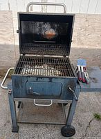 Open charcoal combo smoker grill with cooking grates and grilling tools on side shelf, revealing interior rack and signs of heavy use.
