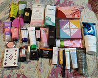 Photo showing entire makeup lot on a patterned quilt including skincare tubes, eyeshadow palettes, lip products, powders, and assorted beauty items