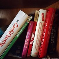 Seven cookbooks and one birthday numerology book arranged upright in a shelf, titles visible