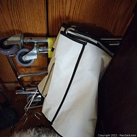 Image showing the laundry rack components folded and leaning against a wooden wall. Visible caster wheels and white fabric with black trim.