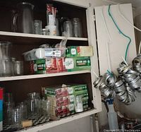 Shelf holding multiple unopened boxes of Ball Mason jars, assorted empty jars, and metal jar rings hanging on side.