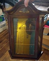 Wooden wall mounted jewelry box with glass front door and carved shell motif on top, showing three glass shelves inside.