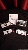 Four Kodak Instamatic cameras arranged on black cloth against red curtain background with lot number 46 sign.