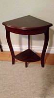Front angle of a dark brown wooden corner table with curved legs and a triangular bottom shelf, placed against a wall on a carpeted floor.