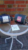 Photo featuring two electric motors on a round table with a brick wall background, one motor blue and the other gray, both visibly labeled.