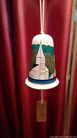 Full view of the white wind bell hanging with handpainted church scene and background trees.