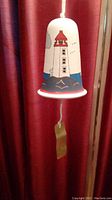 Full view of the white clay wind bell with lighthouse design, hanging by a white rope with a visible clapper at the bottom.