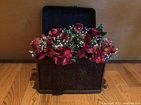 Frontal view of hinged wooden chest with woven pattern open and filled with silk red roses and white baby's breath flowers on wooden floor.