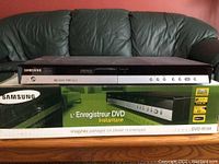 Samsung DVD-R150 DVD recorder placed on top of the original retail box with a leather couch background.