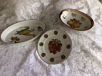 Three ceramic Royal Worcester serving dishes on lace fabric: oval sole dish with corn motif, round plate with fruit design, and oval serving bowl with fruits.