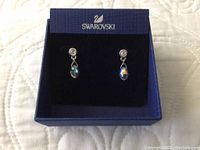Pair of Swarovski drop earrings displayed in original blue Swarovski box on a quilted white surface.