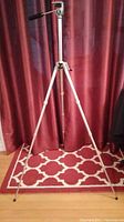 Full view of the extended silver tripod standing on a patterned rug with a red curtain background.