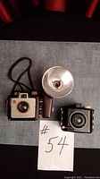 Both Kodak Brownie cameras and the external flash unit together shown on a fabric surface with a tags #54