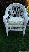 Front view of white wicker chair with green and white patterned cushion, arched back and rounded armrests on grass
