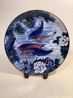 Full view of circular charger plate on display stand. Shows the koi fish swimming motif with white flowers and green pine branch accents. Colors predominantly blue, green, orange, and white.