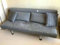 Gray fold down futon sofa showing four pillows: two smooth rectangular gray cushions and two textured darker gray patterned pillows. Cushion shows some stains.