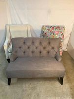 Three seating pieces: small grey tufted loveseat, white swoop-arm armchair, and floral armchair in a room with plain white backdrop.