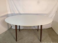 Oval dining table with matte white top and dark walnut legs shown against white backdrop and concrete floor, visible from front angle.
