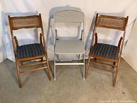 Front view of three folding chairs: two identical wooden folding chairs with caned backs and striped seat cushions flank a gray metal folding chair with padded seat and back.