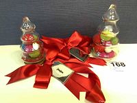 Two glass pine trees filled with colorful felt pompoms, two red gift ribbons with metal Neiman Marcus tags laid out on a white surface.