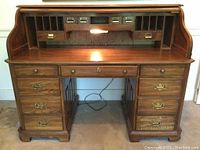 Front view of the roll top desk with upper hutch cubicles and drawers, and lower seven drawers visible. Desk lamp is switched on illuminating the writing surface.