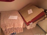 Stack of Pottery Barn placemats in several warm colors and stripes, with a crocheted afghan and woven throw blanket next to them