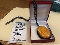 Full view of the Baltic amber pendant on a metal chain displayed in an open jewelry box and a tag showing "Amber Pendant, Metal Chain".