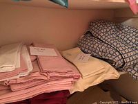 Folded bed linens and comforter arranged on a shelf including pink, white, cream linens and blue patterned comforter
