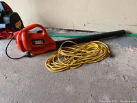 Full side view of the orange Black & Decker leaf blower laying on the ground with yellow extension cord coiled next to it.