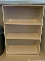 Front view of white MDF bookcase showing empty rectangular frame with two shelves inside, peg holes visible along side panels for shelf height adjustment.