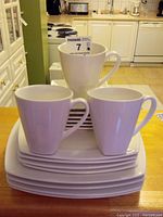 Stack of 3 white mugs on top of stacked square plates showing handles and shape.