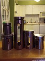Four Oggi brand cylindrical canisters with dark stainless steel finish and glass windows, arranged by size on a brown wooden table in a kitchen setting.