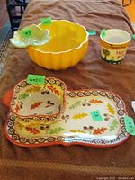 Yellow ceramic chip and dip bowl with attached leaf-shaped dip dish, autumn-themed chip and dip tray set with fall leaves pattern, and small ceramic planter with floral text sitting on fabric surface.