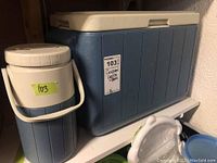 Blue Coleman cooler model 5277 with beige lid and matching blue and beige Coleman jug on a shelf.