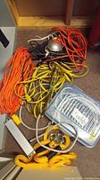 Multiple coiled cords in orange, yellow, and black along with yellow braided rope, power bar inside plastic tote, and halogen work light with protective cage.