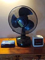 Front view of Windmere table fan centered with Sylvania and Venturer clock radios on either side on wooden surface.