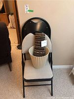 Photo showing a folding chair with beige upholstered seat and backrest, holding a striped floor mat and a white plastic waste basket.