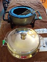 Rival electric fondue pot with forks sitting on graph-patterned surface; silver plated serving dish with lid and glass insert in front