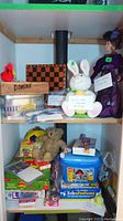 Two wooden shelves filled with a variety of games and toys. Top shelf contains a chess board, domino box, plush bunny, and purple doll. Bottom shelf includes a teddy bear, soccer ball, Telestrations game, Monopoly, Clue, and other miscellaneous games and toys.