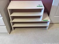 Two white MDF laminate stackable shoe storage shelves of different sizes on carpeted floor, positioned side-by-side near a door and wall.