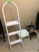 Photo showing the full view of the white metal step ladder with three steps, white paint worn with rust and a white plastic stool next to it plus two additional small step stools.
