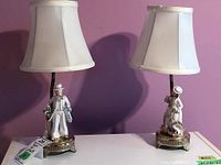 Front view of pair of porcelain figurine table lamps with shades, showing detailed Victorian-style male and female figures on bases.