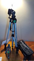 Photo showing a silver Soligor tripod extended and standing on a wooden surface, accompanied by a black Soligor carrying case and a flexible mini tripod with blue joints.