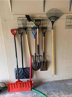 Photo shows a collection of yard tools hanging on a garage wall including red snow shovel, metal spades, garden fork, and leaf rake with yellow handle.
