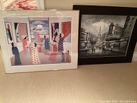 Photo showing both framed art prints leaning against a wall on a carpeted floor; John Lim signed print is colorful with figures and the Parisienne scene is black and white.