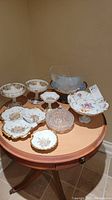 Display of hand-painted china serveware including footed bowls and multi-sectioned dish with gold floral patterns plus crystal candy dish on round wooden table