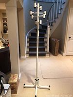 Full view of the coat rack showing the vertical pole with multiple arms and black end caps, placed in a hallway with carpet flooring and staircase in the background.