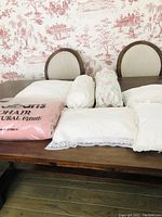 Photo showing multiple cushions of varying shapes and linens along with a packaged pink mohair blanket on a wooden table with two wooden chairs behind.